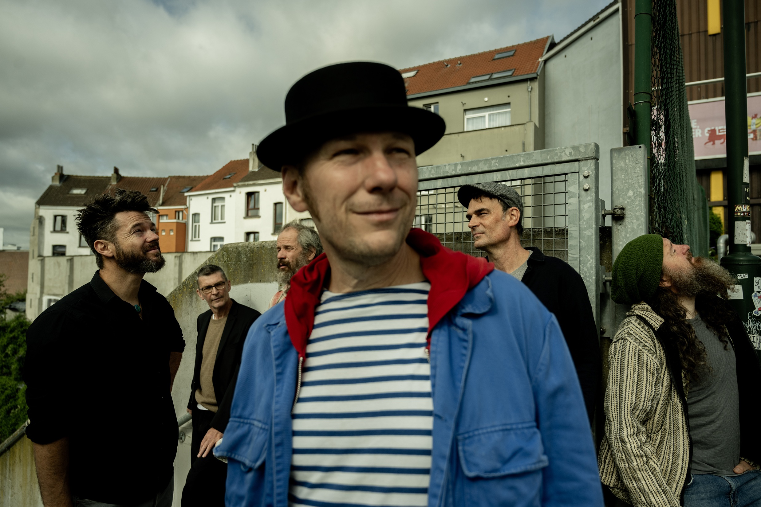 Eine Gruppe Männer, im Vordergrund ein lächelnder Mann mit Hut, offener Jacke und gestreiftem Shirt